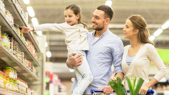 Ein kleines Mädchen zeigt auf ein Produkt im Supermarkt, während der Vater sie hoch hält.